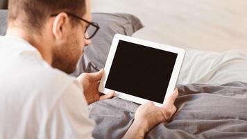 Man Using Digital Tablet With Empty Screen Browsing Internet Lying In Bed In Bedroom At Home. Computer Blank Display For Application And Website Advertisement Concept. Panorama, Mockup photo