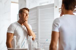 Young man touching his face with hand and gazing confidently while standing in front of the mirror photo