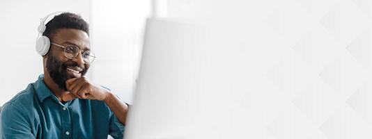 A young African American entrepreneur is working in an office setting, wearing headphones and smiling as he engages with his tasks on a computer. The atmosphere is focused and productive, copy space photo