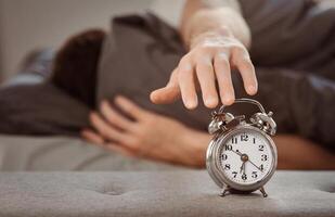 Time To Get Up. Man Turning Off Alarm-Clock Waking Up Wrapped In Blanket Lying In Bed At Home. Shallow Depth photo