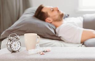 Sleeping Pills Lying Near Alarm-Clock While Asleep Man Sleeps In Bed Resting In Bedroom At Home. Insomnia Treatment, Medication And Medicine For Sleeplessness. Shallow Depth, Selective Focus photo