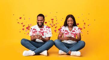 A couple sits together on a bright yellow background, each focused on their smartphones. They are sharing affectionate messages for Valentine's Day, surrounded by heart motifs. photo