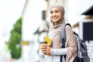 Smiling muslim girl student drinking coffee on street before studying, copy space photo