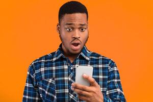 Phone Problem. Shocked African Guy Looking At Smartphone Reading Message With Shocking News Standing Over Yellow Background In Studio photo