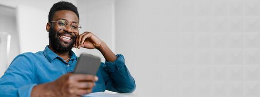 A young African American manager is smiling while using his smartphone at the office. He is engaged and relaxed, showcasing success and a modern lifestyle in a professional environment, copy space photo