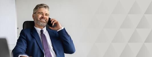 Successful Mature Businessman In Suit Talking On Mobile Phone At Office, Handsome Middle Aged Man Entrepreneur Sitting At Desk With Laptop And Enjoying Pleasant Call With Business Partner, copy space photo