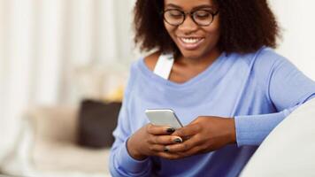 Scrolling News Feed. Closeup of african american smiling lady holding mobile phone, typing sms message, sitting on couch. Communication Concept. Black woman browsing internet, surfing web, using app photo