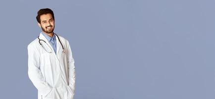 Portrait of confident Indian medical doctor in lab coat looking at camera on violet background, panorama photo