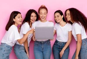 Group Of Multiethnic Ladies Using Laptop Computer Reading Female Blog Together Standing Over Pink Studio Background. Women Browsing Internet. E-Learning, Online Education And Technologies photo