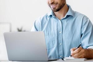 Millennial male entrepreneur using laptop and taking notes at workplace in modern office, looking at computer screen and writing something to notepad, sitting at desk, planning budget, free space photo