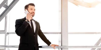 Cheerful rich businessman standing in airport, talking on mobile phone with business partners and looking through the window at airplane, having business trip, panorama with copy space, sun flare photo