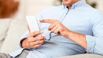 Scrolling News Feed. Closeup of unrecognizable smiling guy holding mobile phone, typing sms message, sitting on the couch. Communication Concept. Adult man browsing internet, surfing web, using app photo