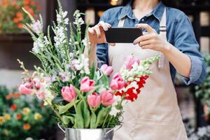 Florist at work. Woman in apron and protective mask makes photo on smartphone of bouquet in flower shop, copy space