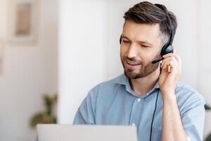 Hotline Operator. Portrait Of Handsome Male Call-Center Employee Wearing Headset At Workplace In Office, Consulting Client, Sitting At Desk Using Laptop Computer For Customer Support, Free Space photo