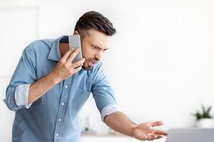 Problems At Work. Stressed Angry Businessman Emotionally Talking On Cellphone, Having Troubles With Laptop Program, Speaking With Customer Support, Displeased Man Looking At Computer Screen photo