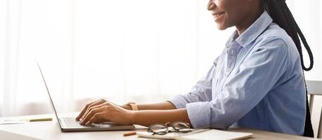 Smiling African American Freelancer Woman Working On Laptop Computer At Home Office, Sitting At Desk Near Window, Self-Employed Lady Typing On Keyboard, Enjoying Remote Job, Side View With Copy Space photo