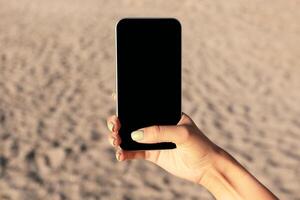Female Hand Holding Mobile Phone With Blank Screen For Your Text In Front Of Beach Sand Outside. Panorama, Mockup, Cropped photo