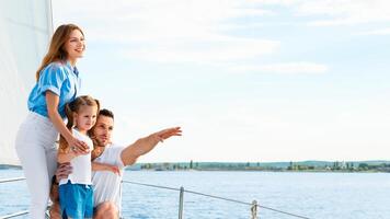 Sailing. Happy Family Embracing Standing On Yacht Looking And Pointing Aside At Sea Water Outdoors. Parents And Daughter Relaxing On Sailboat Enjoying Summer Weekend Together. Panorama, Copy Space photo