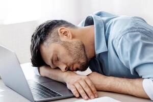 Sleep At Work. Exhausted overworked man employee napping at workplace in modern office, tired after working with laptop and reports, sitting at desk, placed head on hands, having job fatigue photo