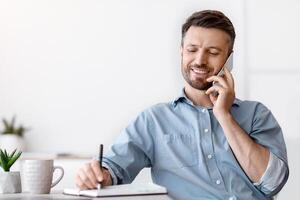 Handsome Young Businessman Talking On Cellphone And Taking Notes While Working In Office, Holding Pen And Writing In Notepad, Male Entrepreneur Sitting At Desk And Enjoying Phone Call, Free Space photo