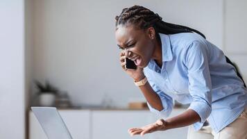 Problems At Work. Stressed Black Businesswoman Talking On Cellphone And Screaming, Having Troubles With Laptop Program, Free Space photo