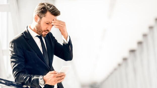 Tired businessman standing by window in airport terminal, using mobile phone, touching his forehead, having headache, copy space. Entrepreneur having business problems, reading emails on smartphone photo