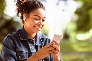 Happy Black Lady Using Smartphone Phone Texting In Social Media Chat App Sitting In Park Outdoor. Online Texting And Messaging, Mobile Applications For Cellphone Concept photo