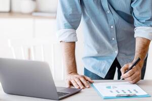 Cropped shot of businessman taking notes to financial report, working with documents, unrecognizable male entrepreneur holding pen in hand, signing contract while standing at desk in modern office, photo