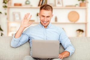 Cheerful guy making call, sitting with computer on his lap, waving with hand, wearing earbuds photo