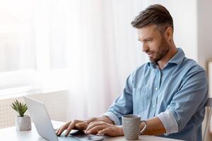 Unemployed man scrolling job search website at home using laptop computer, handsome male freelancer working remotely at desk near window, typing on keyboard, answering emails, copy space photo