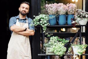 Confident businessman, flower shop owner and ad of modern studio in rustic style. Smiling millennial confident male hipster in apron with crossed arms at store with plants in pots and vases outdoor photo