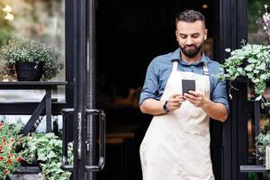 Online blog about flowers, work of florist in modern studio and ordering bouquet. Cheerful young bearded european man in apron typing on smartphone at front door of rustic shop with different plants photo