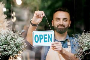 New rules for store opening after covid-19 quarantine and positive emotions of owner. Happy young attractive bearded guy in apron turns sign with inscription is open on door in flower studio, outdoor photo