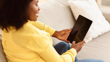 Unrecognizable black girl using digital tablet sitting on couch. Back shot, blank screen of computer for copy space photo