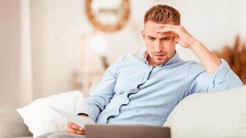 Foused man working from home, sitting on sofa, holding document and looking at personal computer photo