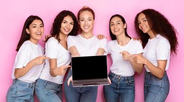 Five multiracial ladies showing laptop with empty computer screen recommending internet website standing posing over pink studio background, smiling to camera. Computers and online services. Mockup photo