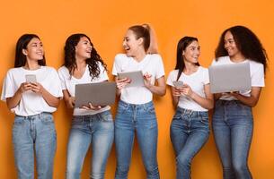 Multiethnic Women Using Gadgets, Computers And Smartphones Browsing Internet And Networking In Social Media Online Standing Over Yellow Studio Background. Internet, Technology And Modern Lifestyle photo
