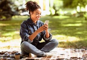 Cheerful African American Lady Using Smartphone Texting In Social Media Mobile App Sitting In Park Outside. Online Communication Applications Concept. Free Space For Text photo