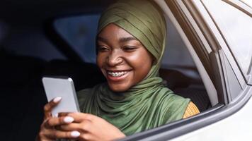 Smiling Black Muslim Lady In Hijab Using Smartphone While Riding Car On Backseat, Browsing Internet Or Texting With Friends, African Woman Enjoying Online Communication, Closeup Shot Through Window photo