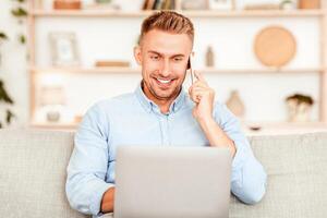 Corporate Communication. Portrait of smiling young entrepreneur making phone call, typing on laptop, sitting on the couch in living room. Confident adult using computer for work, consult client online photo