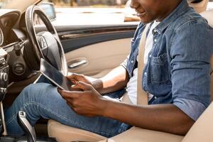 Positive black guy using digital tablet while driving car, setting destination to go, using tracking mobile app, side view, copy space, sun flare. Afro american driver checking gps on his pad photo