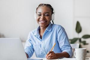 Young Smiling Black Female Student Studying Online With Laptop, Listening Music And Taking Notes, Sitting At Desk At Home, Enjoying Distance Learning, Preparing For Exams With Computer, Copy Space photo