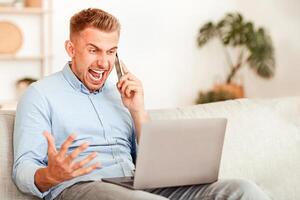 Angry irritated guy talking on mobile phone and shouting, sitting on sofa, using laptop, copy space photo