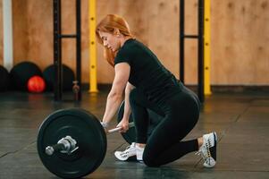 View from the side. Sitting with barbell on the floor. Beautiful strong woman is in the gym photo
