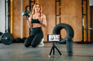 Smartphone is on the tripod, live ,stream,, capturing . Beautiful strong woman is in the gym photo