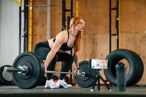 Side view, with barbell. Smartphone on tripod capturing . Beautiful strong woman is in the gym photo