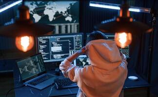 Sitting,, in white hoodie. Young professional female hacker is indoors by computer with lot of information on displays photo