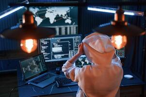 View, from behind. Young professional female hacker is indoors by computer with lot of information on displays photo