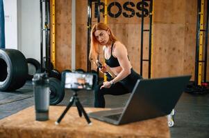 Laptop,, bottle and smartphone on tripod is on the table. Beautiful strong woman is in the gym photo