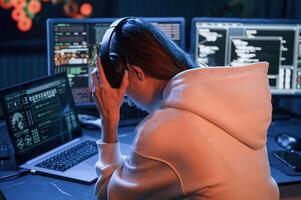 Taking of headphones. Young ,professional female hacker is indoors by computer with lot of information on displays photo
